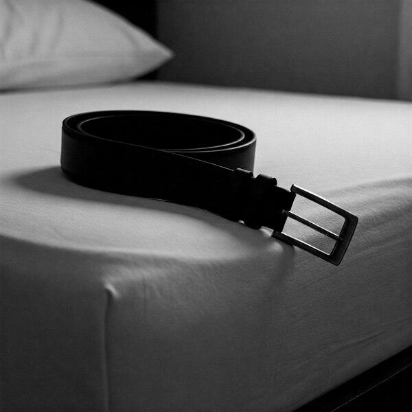 A black leather belt on a sterile white mattress, evoking ritual degradation, object use, and non-romantic domination.